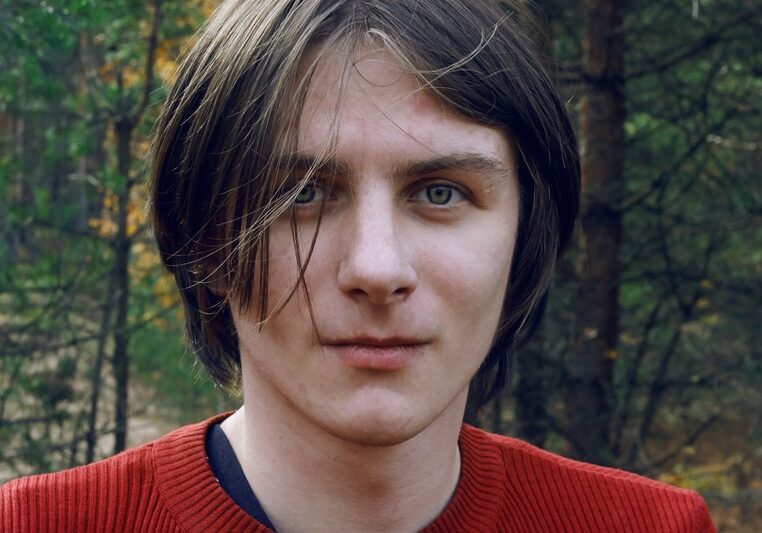 A handsome guy with long tousled hair and green eyes stands in the middle of the autumn forest