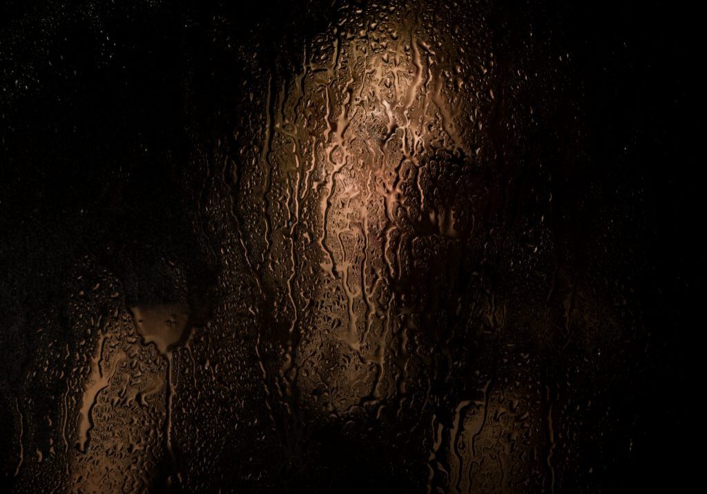 Smooth portrait of sexy model, posing behind transparent glass covered by water drops. young melancholy and sad woman portrait.